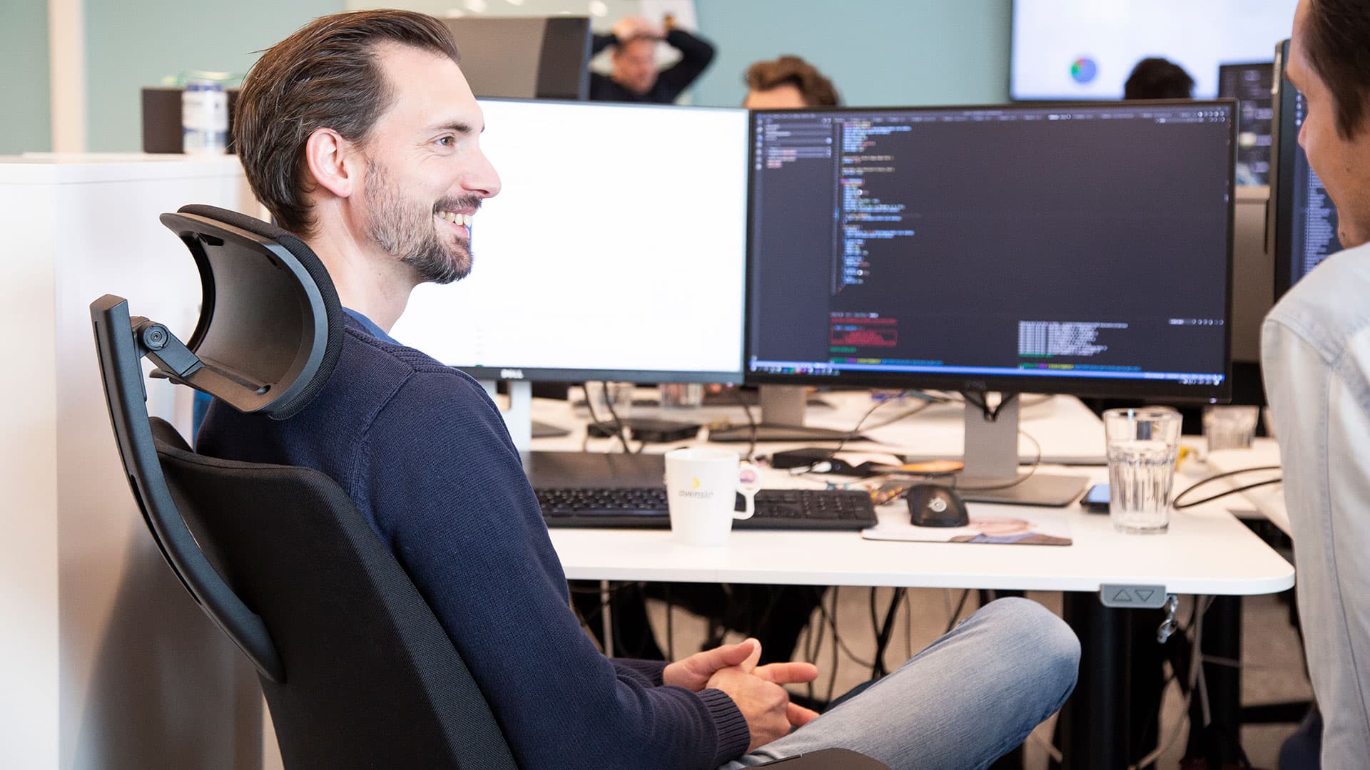 En som sitter bakåtlutad i sin kontorsstol och pratar med en kollega bredvid sig